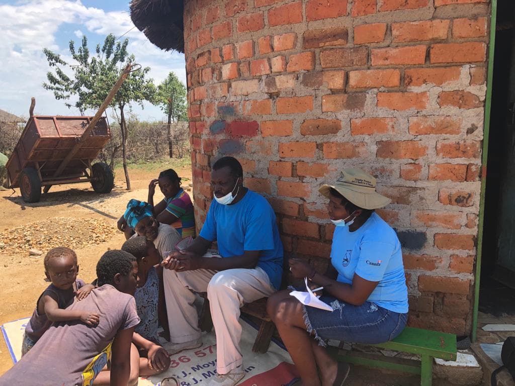 Community members seated outside a rural homestead in conversation with Trinity Project field staff.